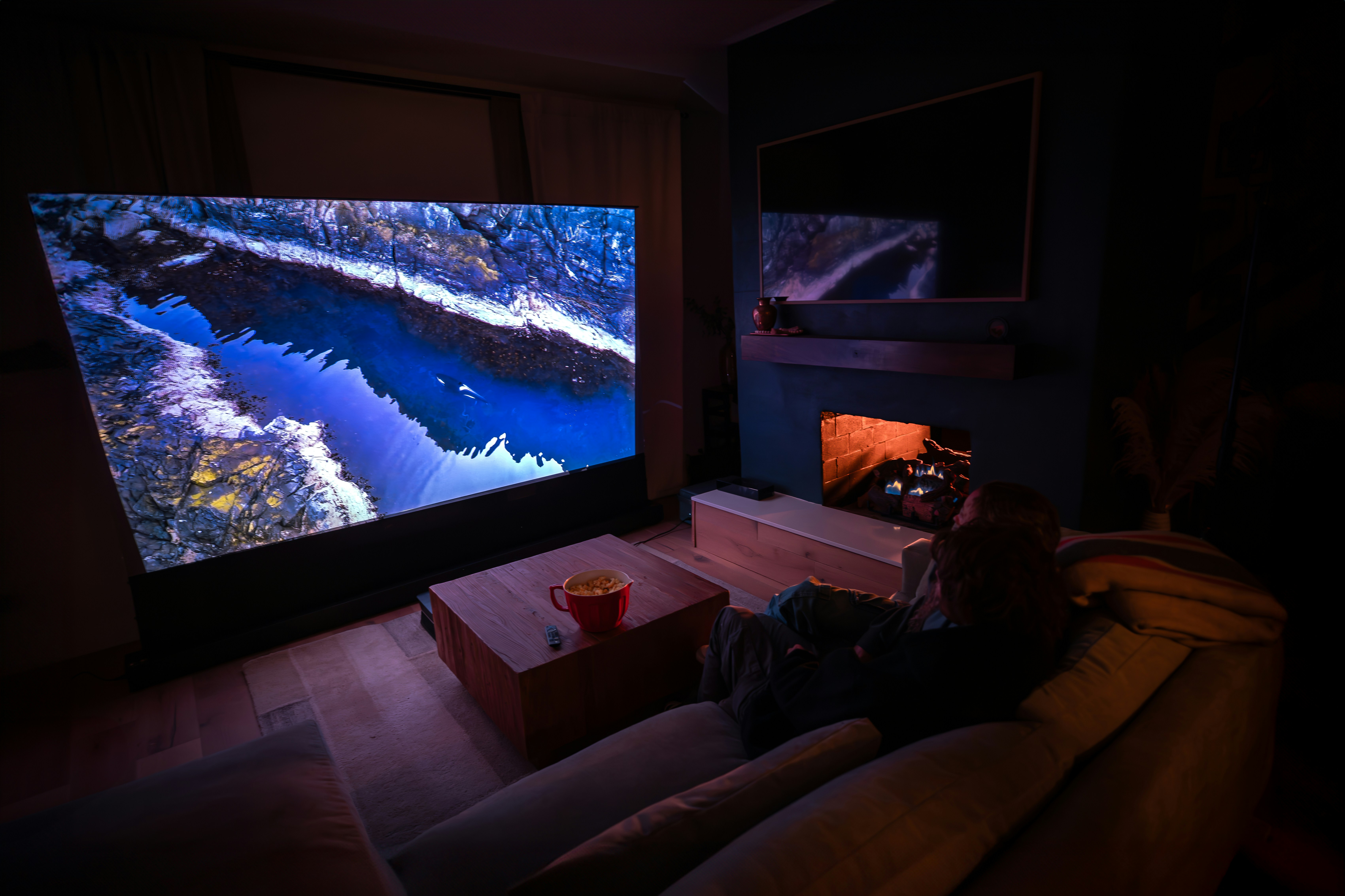 Two people sitting on a couch watching nature river landscape on AWOL Vision 4K UST projector. by TV Brands A person sitting on a couch in front of a flat screen TV